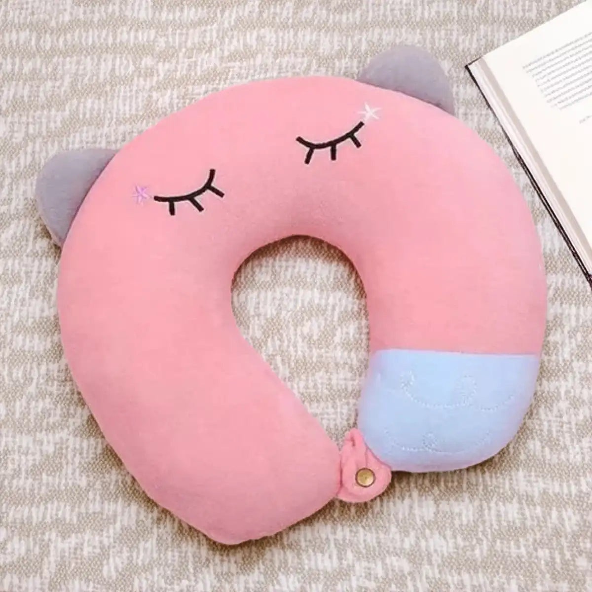 Almohada de cuello color rosa con diseño de gatito kawaii obre una mantita y cerca a un libro