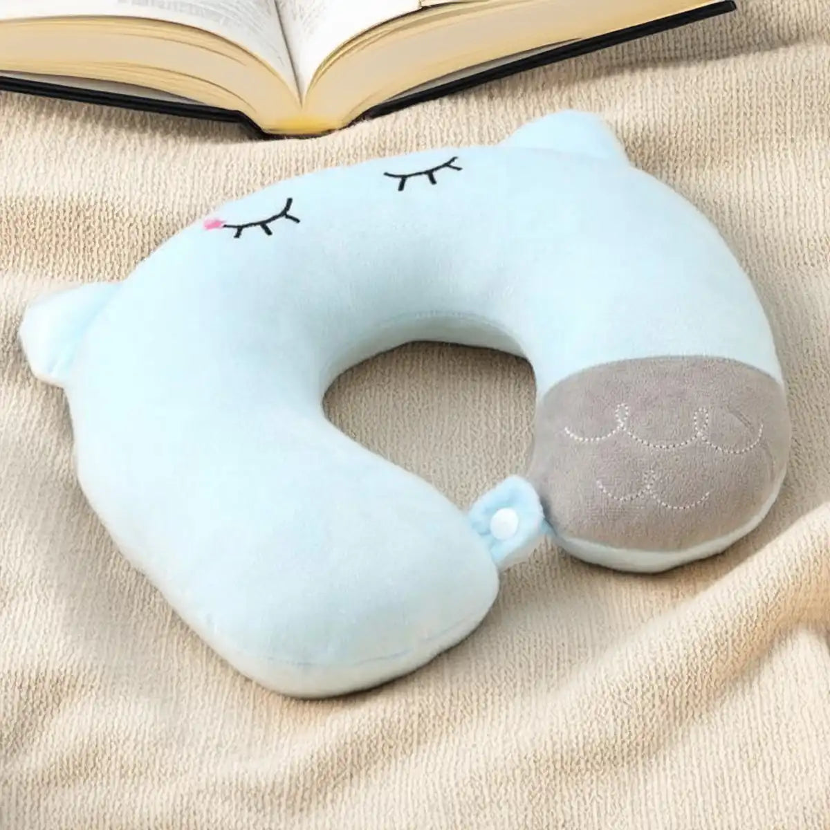 Almohada de cuello color azul bebé con diseño de gatito kawaiis obre una mantita y cerca a un libro